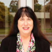 A headshot of Professor of Film and Media Studies, Dick Wolf Director of the Carsey-Wolf Center, and Presidential Chair in Media Studies, Patrice Petro. The image depicts a woman with black hair, wearing a black blazer, with a floral button up shirt. She is smiling and posed in front of a set of doors.