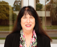 A headshot of Professor of Film and Media Studies, Dick Wolf Director of the Carsey-Wolf Center, and Presidential Chair in Media Studies, Patrice Petro. The image depicts a woman with black hair, wearing a black blazer, with a floral button up shirt. She is smiling and posed in front of a set of doors.