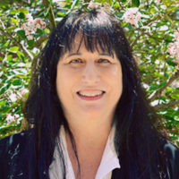 Patrice Petro, the Dick Wolf Director of the Carsey-Wolf Center, appears against a background of green leaves and pink flowers. She has long black hair and is smiling.