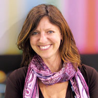An image of Professor Jennifer Holt against a multicolored striped background. Professor holt has reddish brown shoulder length hair. She is wearing a pink scarf, and is smiling.
