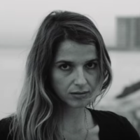 Actor/producer Lynn Cruz appears againsta seascape with a building in the background. She has light brown hair and is looking at the camera with a serious expression.