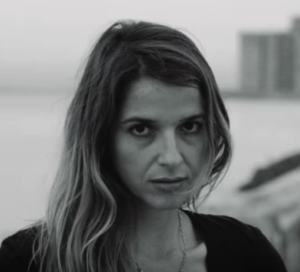 Actor/producer Lynn Cruz appears againsta seascape with a building in the background. She has light brown hair and is looking at the camera with a serious expression.