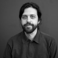 Director Miguel Coyula appears against a neutral grey background. He has long black hair in a ponytail, is wearing a dark shirt, and has a dark beard and mustache.