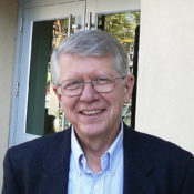 This is a headshot of professor Charles Wolfe. The image depicts a man with glasses, and blue blazer, and a pin stripe blue button up shirt. He is smiling towards the camera, posed in front of a set of doors.