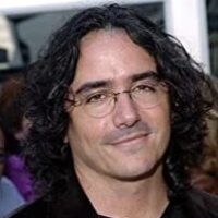 Headshot of director Brad Silberling. It depicts a man with shoulder length hair in glasses, a black button up shirt, and a black blazer. He is smiling and appears to be posed in front of a crowd of people.