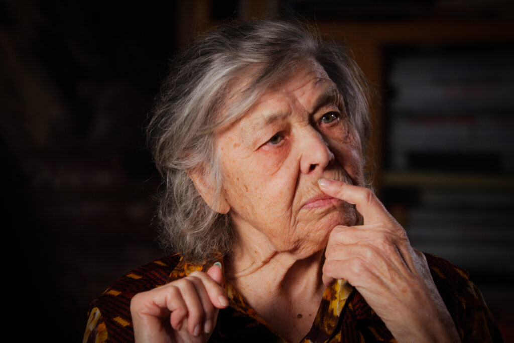 Production still from the film, Women of the Gulag. An elderly woman sits with a finger across her lips