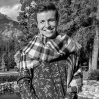 Headshot of Tyler Morgenstern. The black and white image depicts a man wearing a scarf and floral button-up long sleeve shirt. He is smiling and posed in front of a nature background which features a pond, large trees and mountains.