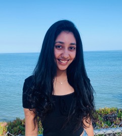 A headshot of communication student Anika Walia. She is smiling at the camera. She has black hair and is wearing jeans and a black shirt with a neck pendant. She stands against a fence on the cliff that looks out to the ocean.