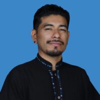 This is a headshot of Assistant Professor in the Department of Chicana and Chicano Studies at UCSB, Giovanni Batz. The image depicts a man posed in front of a neutral blue background, he has a goatee, and black stud earrings. He wears a black shirt with embroidered blue lining accents, and a rounded collar.