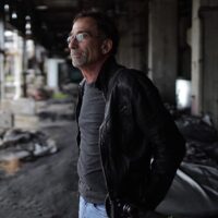 JamesHaymanHeadshot Director James Hayman is shown standing in an urban area facing to the left side of the frame. He has dark hair and glasses, and is wearing a grey tshirt and black jacket.