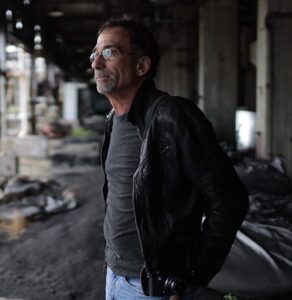 JamesHaymanHeadshot Director James Hayman is shown standing in an urban area facing to the left side of the frame. He has dark hair and glasses, and is wearing a grey tshirt and black jacket.