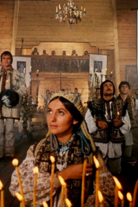 A still from the film Shadows of Forgotten Ancestors, depicting a woman in ornate traditional Ukrainian grab in the foreground behind a row of candles, with a group of men in the background.
