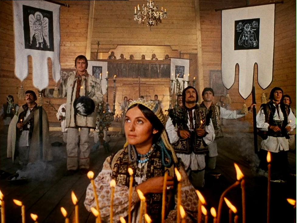 A still from the film Shadows of Forgotten Ancestors, depicting a woman in ornate traditional Ukrainian grab in the foreground behind a row of candles, with a group of men in the background.