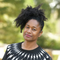 Cathy Thomas, a professor of English at UCSB, is pictured on a leafy green background. She has black curly hair and is smiling with her head tilted to the side. She is wearing a black sweater with white patterns on it.