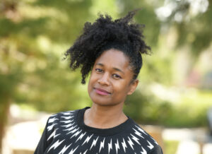 CathyThomas_Headshot Cathy Thomas, a professor of English at UCSB, is pictured on a leafy green background. She has black curly hair and is smiling with her head tilted to the side. She is wearing a black sweater with white patterns on it.