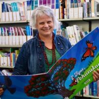 A photo of professor Nicola Daly. She is holding a large, brightly colored children's picturebook and smiling in front of shelves of books. She is wearing a denim jacket and has grey hair.