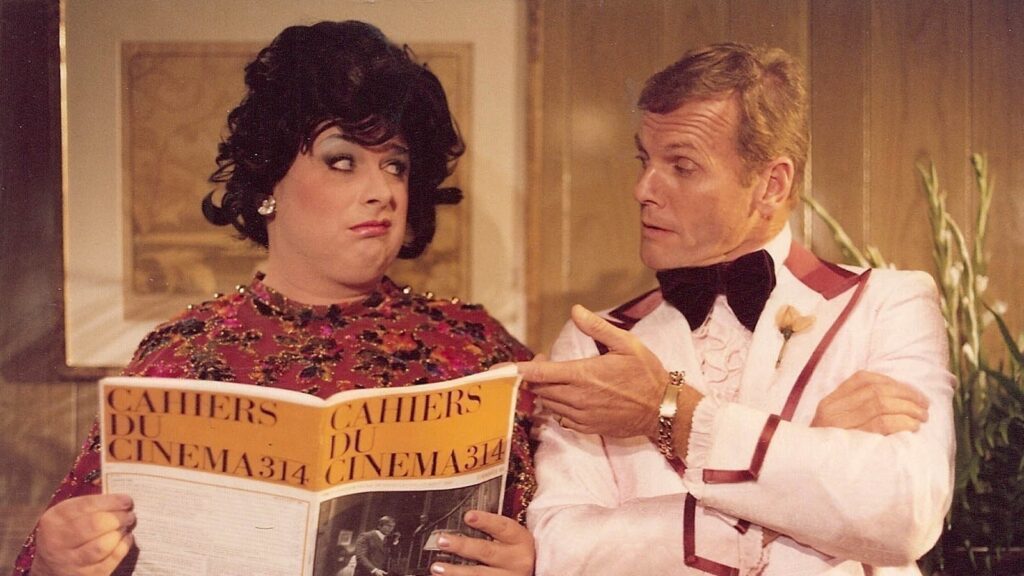 In this still from John Waters' film Polyester, drag queen Divine, dressed in a dark wig and garish dress, and actor Tab Hunter, whose hair is blond and who wears a white uniform, read a copy of the film ciricism journal Cahiers du Cinema.