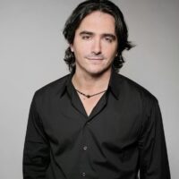 Headshot of director Brad Silberling. It depicts a man with shoulder length hair in a black button up shirt against a neutral background. He is smiling and is wearing a black twine necklace.