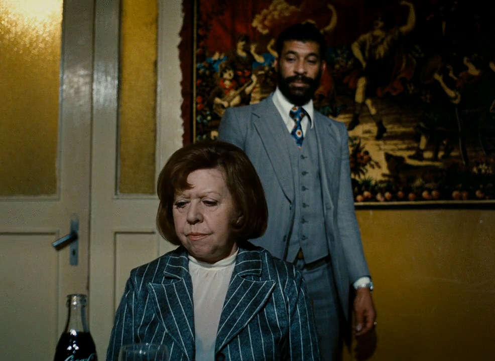 Promotional still for the film Ali: Fear Eats The Soul. The image depicts an older woman in the foreground sitting at a table with a glass soda bottle. She wears a serious, pensive expression, staring at the table in front of her, and a pin stripe blazer. Behind her is the film's namesake, Ali, watching over her shoulder, he wears a grey three piece suit, and has a curious expression.