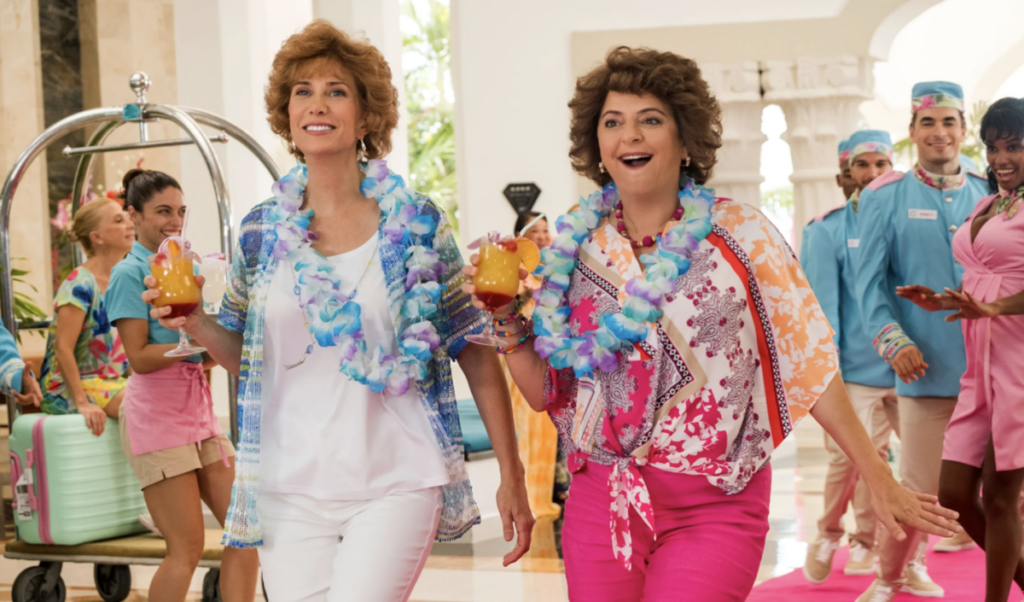 Two women walk through a crowded hotel lobby in bright, tropical clothes, carrying cocktails and smiling.