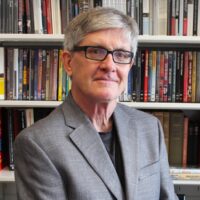 An image of professor Timothy Corrigan. An older man with silver hair and glasses wears a grey suit jacket, sitting in front of a wall of books.