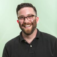 Headshot of writer/comedian Gabe Liedman. He is wears a black polo shirt, half rimmed glasses and he is smiling, posed in front of a neutral green background