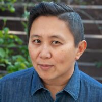 An image of Professor Karen Tongson. She has short dark hair and wears a denim-style, button-up shirt, and is sitting in front of a wooden fence with