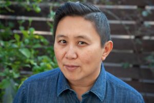 KarenTongson_Headshot An image of Professor Karen Tongson. She has short dark hair and wears a denim-style, button-up shirt, and is sitting in front of a wooden fence with