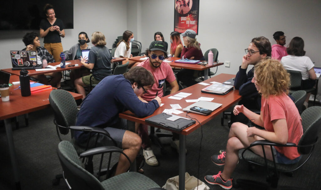 A classroom full of students, gathered in groups around tables, working on screenwriting exercises.