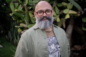 AlejandroFloresAguilar_headshot Headshot of filmmaker Alejandro Flores Aguilar. The image depicts a bearded man, wearing a green button up shirt over a graphic tee. He wears glasses and smiles, posed in front of a a prickly pear cactus.