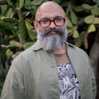 Headshot of filmmaker Alejandro Flores Aguilar. The image depicts a bearded man, wearing a green button up shirt over a graphic tee. He wears glasses and smiles, posed in front of a a prickly pear cactus.