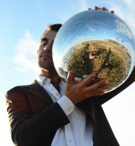 Portrait of Graham Feyl taken from a low angle. He is posing with his eyes closed, and a disco ball held up to half of this face against a sky background. He wears a white dress shirt and grey blazer.