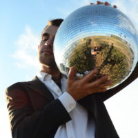 Portrait of Graham Feyl taken from a low angle. He is posing with his eyes closed, and a disco ball held up to half of this face against a sky background. He wears a white dress shirt and grey blazer.