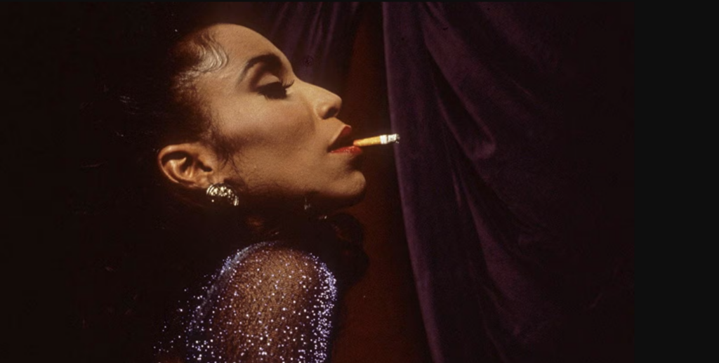 An image from the film Paris is Burning depicting a young drag performer in profile, wearing an evening gown and smoking a cigarette against a black velvet backdrop.