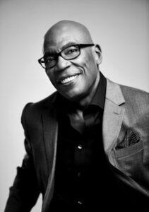 ParisBarclay_Headshot_2023 Black and white headshot of writer/director Paris Barclay. He wears glasses, a three piece grey suit with a black button-up. He is smiling and posed in front of a neutral background.