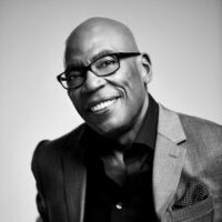 Black and white headshot of writer/director Paris Barclay. He wears glasses, a three piece grey suit with a black button-up. He is smiling and posed in front of a neutral background.