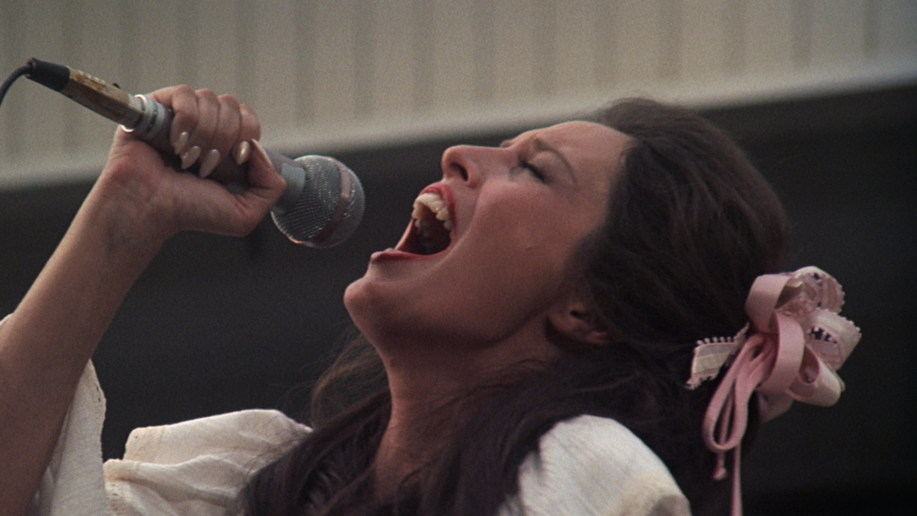 A still from the film Nashville, depicting a young woman with brown hair and a white dress singing passionately into a silver microphone.