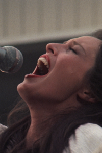 A still from the film Nashville, depicting a young woman with brown hair and a white dress singing passionately into a silver microphone.