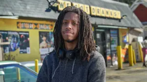 This is a headshot of filmmaker Juel Taylor he wears a grey hoodie and headphones around his neck. He is posed in front of a set for his film They Cloned Tyrone.