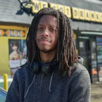 This is a headshot of filmmaker Juel Taylor he wears a grey hoodie and headphones around his neck. He is posed in front of a set for his film They Cloned Tyrone.