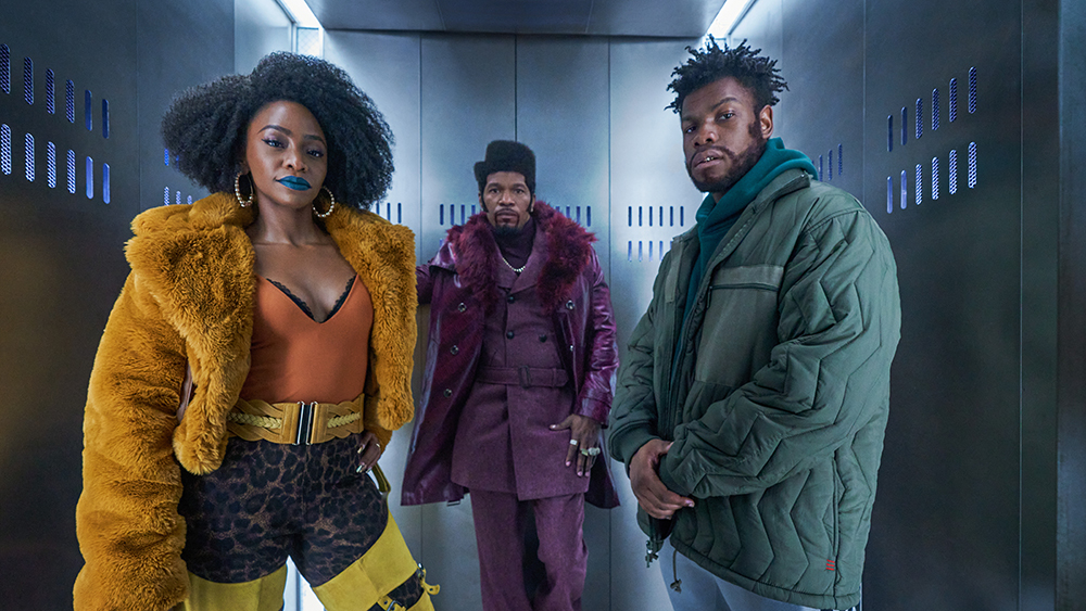 Promotional image for the film They Cloned Tyrone. The image depicts three characters from the film. On the far left is Teyonah Parris as Yo-yo, wearing a monochrome yellow outfit and blue lipstivk. In the center is Jamie Foxx as Slick Charles, he wears a monochrome purple suit and coat, with an asymmetric flat top haircut. On the far right is John Boyega as Fontaine wearing a green hoodie under a green puffer jacket, with grey sweat pants. They are all wearing serious expressions and looking towards the camera.