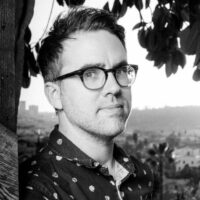 A black and white headshot of the filmmaker Arthur Jones. He wears dark-rimmed glasses, a dark shirt, and is shot against an outdoor landscape.