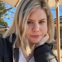 Headshot of graduate student Chelsea Roesch. It depicts a woman with blonde hair and a nose piercing posing in front of an umbrella outside a building.