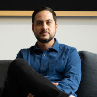 “A portrait of the architectural designer and producer, Giorgio Angelini. It depicts a middle-aged man wearing a blue shirt and black pants. He is posed against a neutral background on a couch with one leg crossed over the other”