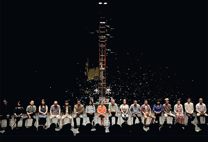 A large group of dancers sits on a darkened stage. A red ladder extends up through the middle of the image.
