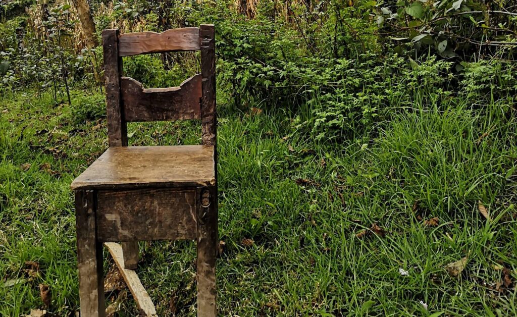 Promotional still for the documentary, Ixiles: Voices from the Shadows of the Past. The image depicts a dilapidated chair in a grassy setting. There is a lot of overgrown grass and trees in the background.