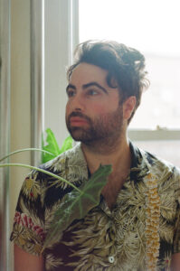 This is a headshot of filmmaker Anthony Banua Simon. The image depicts a man posed in front of a window with a monstera plant leaf in front of him. He is facing away from the camera and posing pensively.