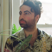 This is a headshot of filmmaker Anthony Banua Simon. The image depicts a man posed in front of a window with a monstera plant leaf in front of him. He is facing away from the camera and posing pensively.