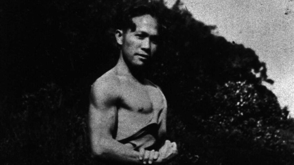 A composite image depicting a black and white photo of a shirtless man, apparently flexing his bicep for the camera, superimposed on an image of a burning sugarcane plantation filled with bright flames. It is a promotional image for the documentary film Cane Fire.