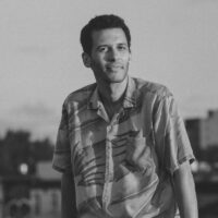 This is a headshot of filmmaker James N. Kienitz Wilkins. The image depicts a man in a graphic button-up, short sleeve shirt. He leans against an object that is out of frame, and he is smiling and posed in front of a blurry city backdrop.